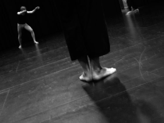 Pieds au sol pendant une session d’entraînement en théâtre physique, avec un autre participant en mouvement à l’arrière-plan.